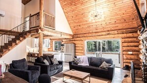 A spacious log cabin interior with a high ceiling, a staircase, a living area with sofas and a coffee table, and a kitchen in the background.