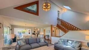 A spacious living area with a large gray sofa, a wooden coffee table, and a staircase leading to an upper level. - Another Day Of Sun - Oceanside 4 BR home w/Hot Tub - 500 feet to beach access (Duck)