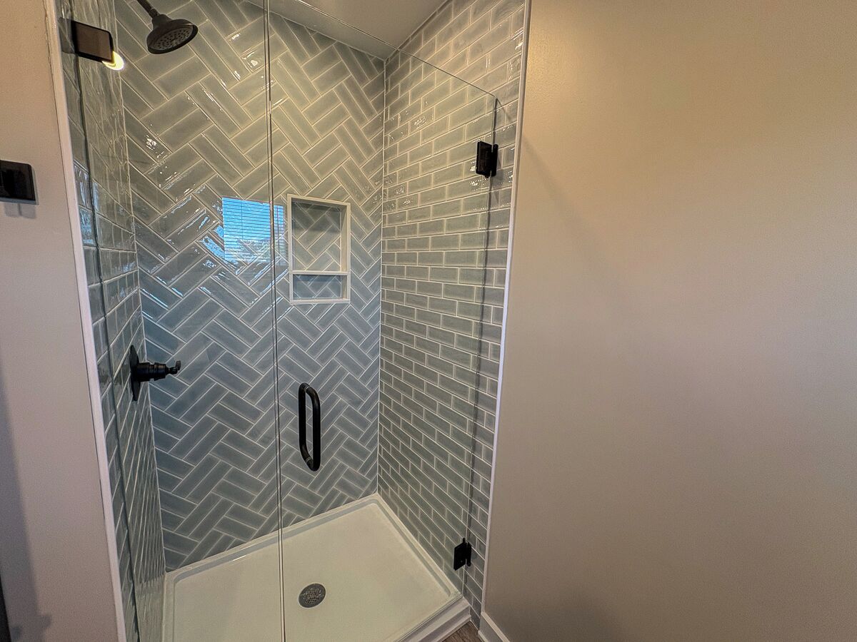 Une douche fermée par des parois de verre, avec un carrelage disposé en motif chevrons, une porte blanche et une poignée noire.