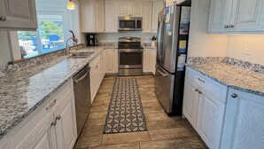 A kitchen with white cabinetry, stainless steel appliances, a patterned rug, and a view of the outside. - Another Day Of Sun - Oceanside 4 BR home w/Hot Tub - 500 feet to beach access (Duck)