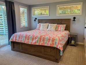 A bedroom with a wooden bed, a beach-themed bedspread, two wall-mounted lamps, and a view of a wooden deck through a window. - Another Day Of Sun - Oceanside 4 BR home w/Hot Tub - 500 feet to beach access (Duck)