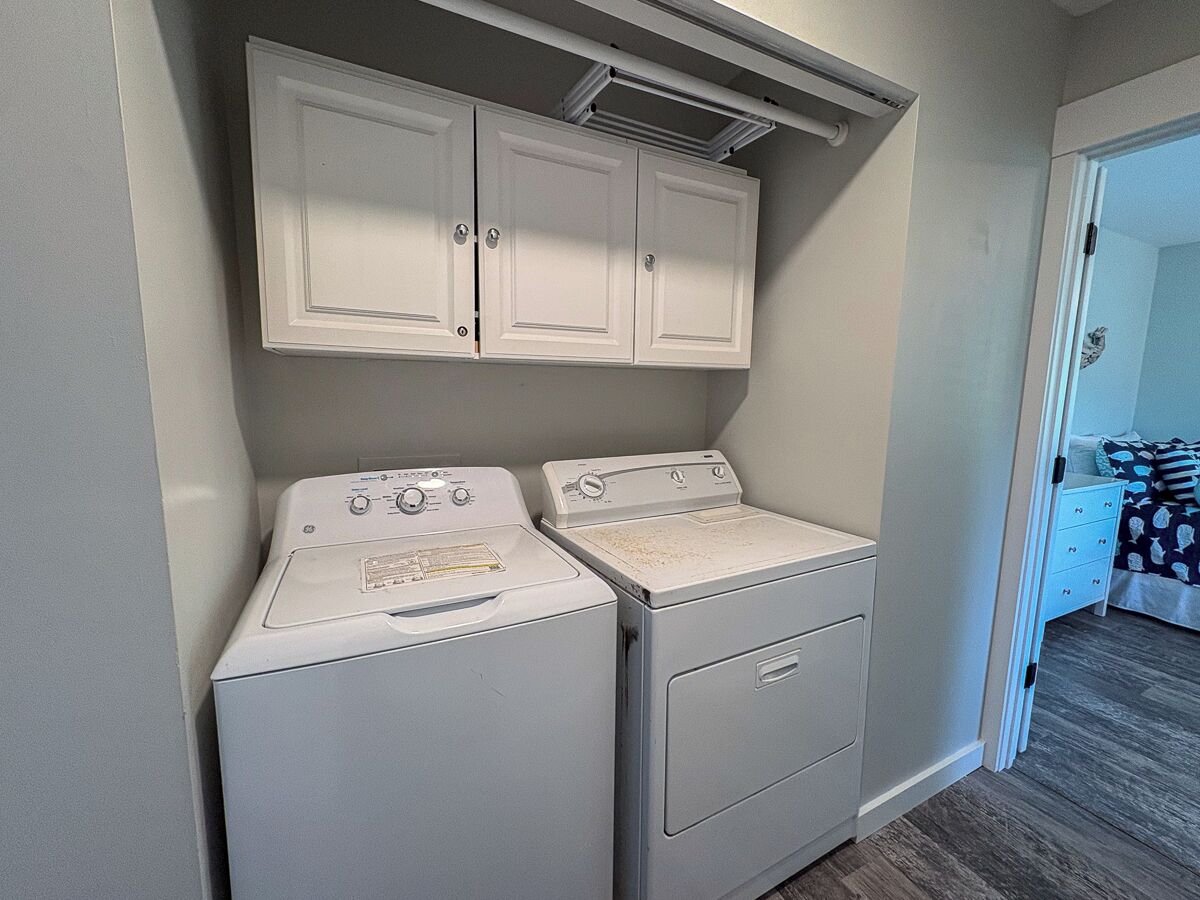 A laundry room with two white washers and dryers, a white cabinet above, and a bed with a striped pillow in the adjacent room.