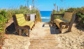 Een houten pad dat leidt naar twee houten banken aan een zandstrand, met een helder uitzicht op de oceaan.