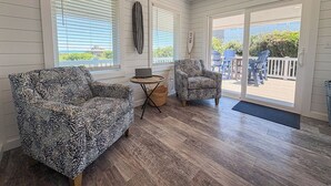 A room with two patterned armchairs, a small round table with a laptop, and a view through a glass door to an outdoor deck. - Another Day Of Sun - Oceanside 4 BR home w/Hot Tub - 500 feet to beach access (Duck)