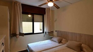 Un dormitorio con dos camas, ventilador de techo, ventana con cortinas, mesita de noche y velador.