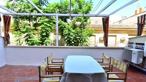 Un patio techado con mesa y sillas, piso de baldosas y vista a un edificio y árboles.