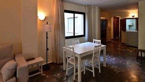 Habitación de hotel con mesa de comedor, sillas, sofá, lámpara, ventana con cortinas y zona de cocina.