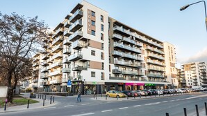 A multi-story residential building with balconies, a pedestrian crossing, and parked cars. - Wola Bloom Apartment (Warszawa)