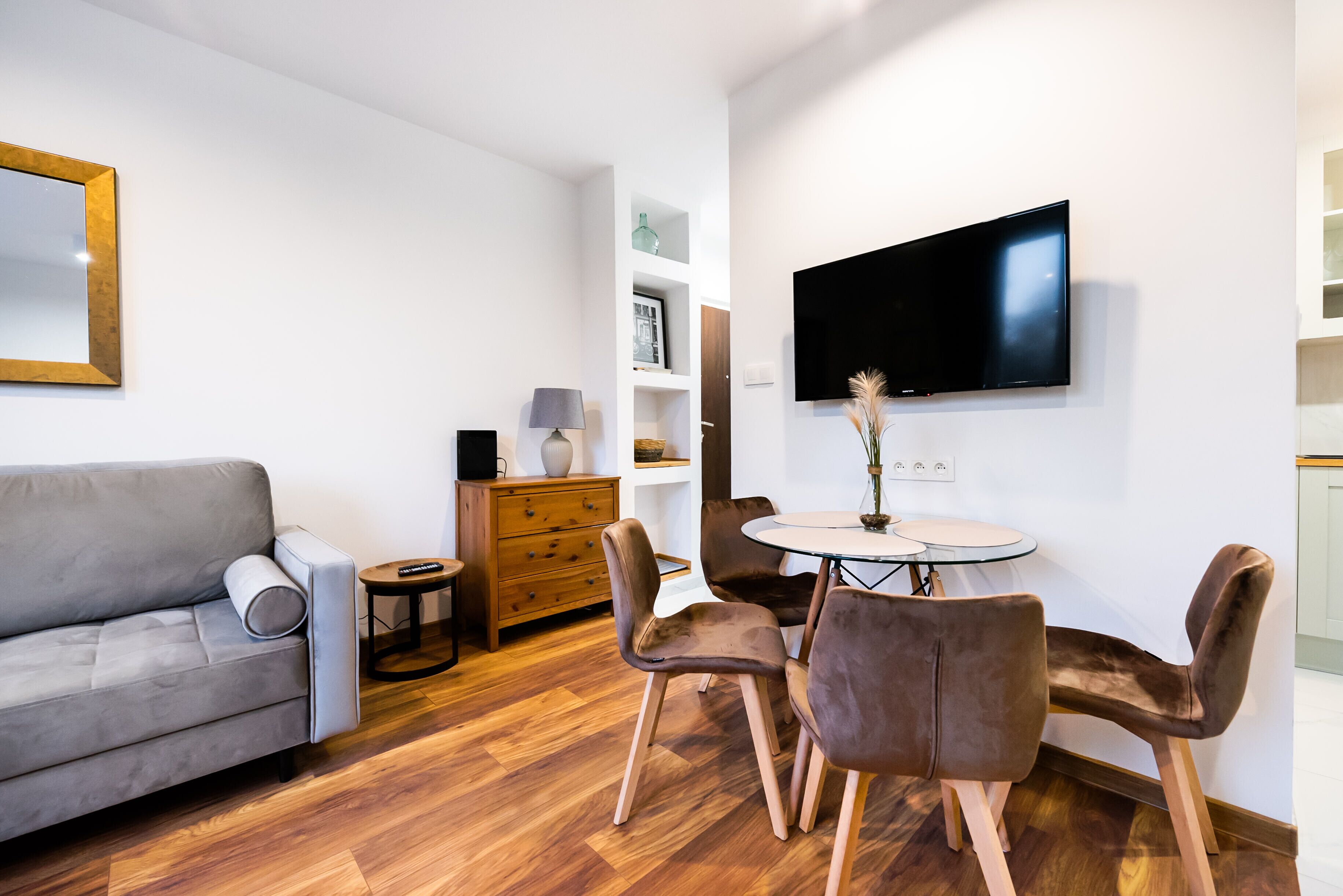 A modern living room with a grey sofa, a round dining table with four chairs, a flat-screen TV mounted on the wall, and a wooden cabinet with drawers.