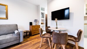 A modern living room with a grey sofa, a round dining table with four chairs, a flat-screen TV mounted on the wall, and a wooden cabinet with drawers. - Żoliborz Elite Apartment (Warszawa)