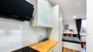 A compact room with a kitchen area featuring a black induction cooktop, white cabinets, and a wooden countertop. There is a bed with a dark-colored mattress and white headboard, and a window with a view of trees. - Żoliborz Elite Apartment (Warszawa)