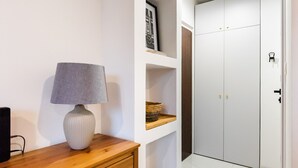 A hallway with a wooden dresser, a lamp, and a white tiled floor leading to a closet with a door. - Żoliborz Elite Apartment (Warszawa)