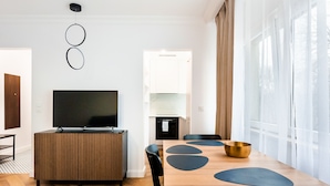 A modern living room with a flat-screen TV, a dining area with a wooden table and chairs, and a kitchen in the background.