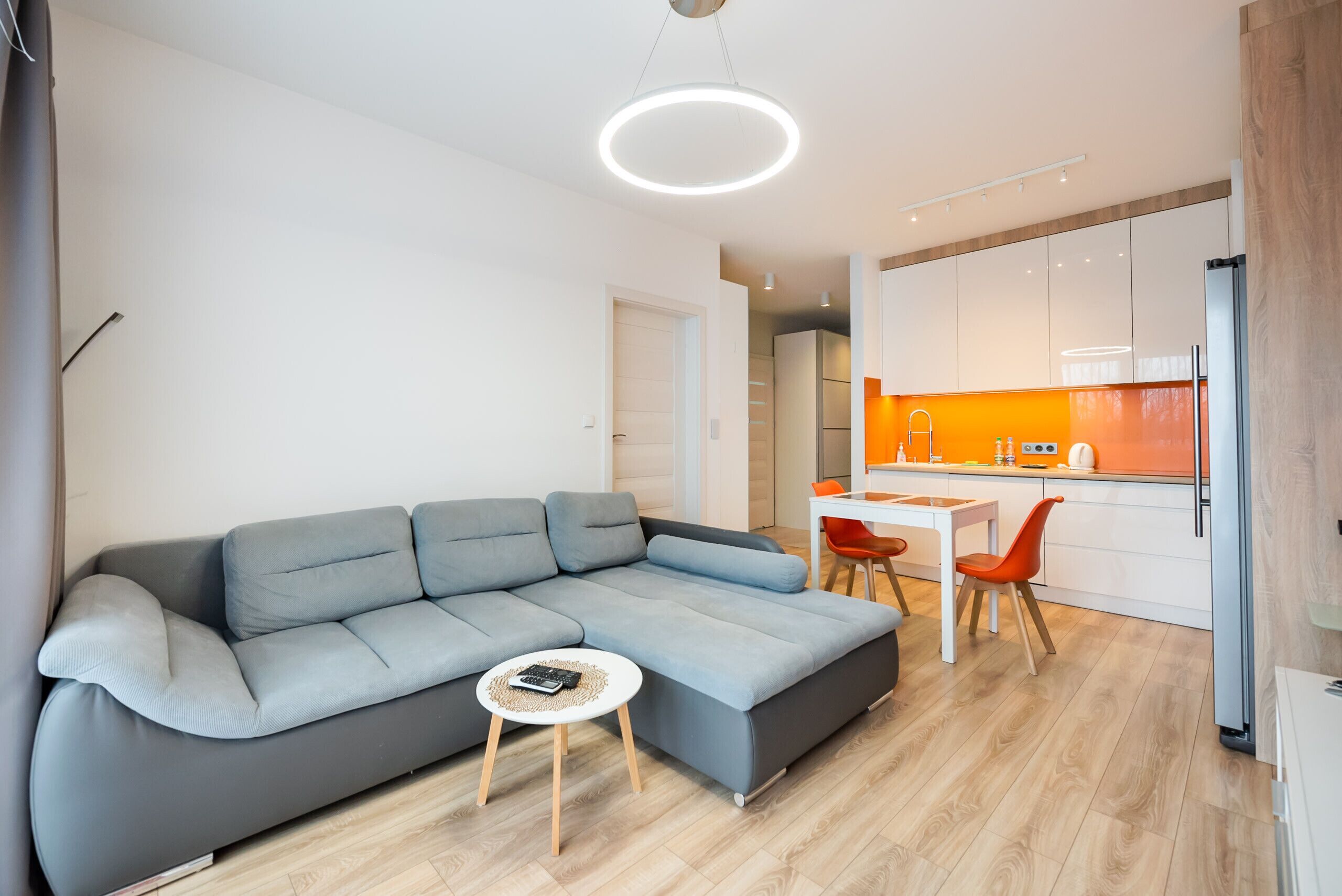 Ein modernes Wohnzimmer mit einem grauen Ecksofa, einem kleinen runden Couchtisch und einer Küchenzeile mit weißen Schränken und orangefarbenen Akzenten.