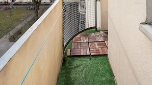 Een smal balkon met een groen kunstgrasveld, een metalen hekwerk en uitzicht op een meerverdieping tellend gebouw op de achtergrond.