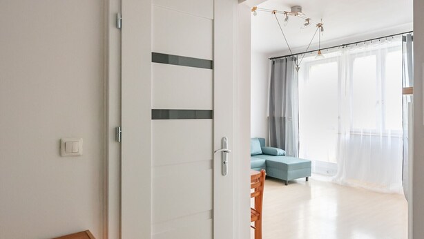 Een kamer met een houten dressoir, een witte deur en uitzicht op een zithoek met een bank en gordijnen.