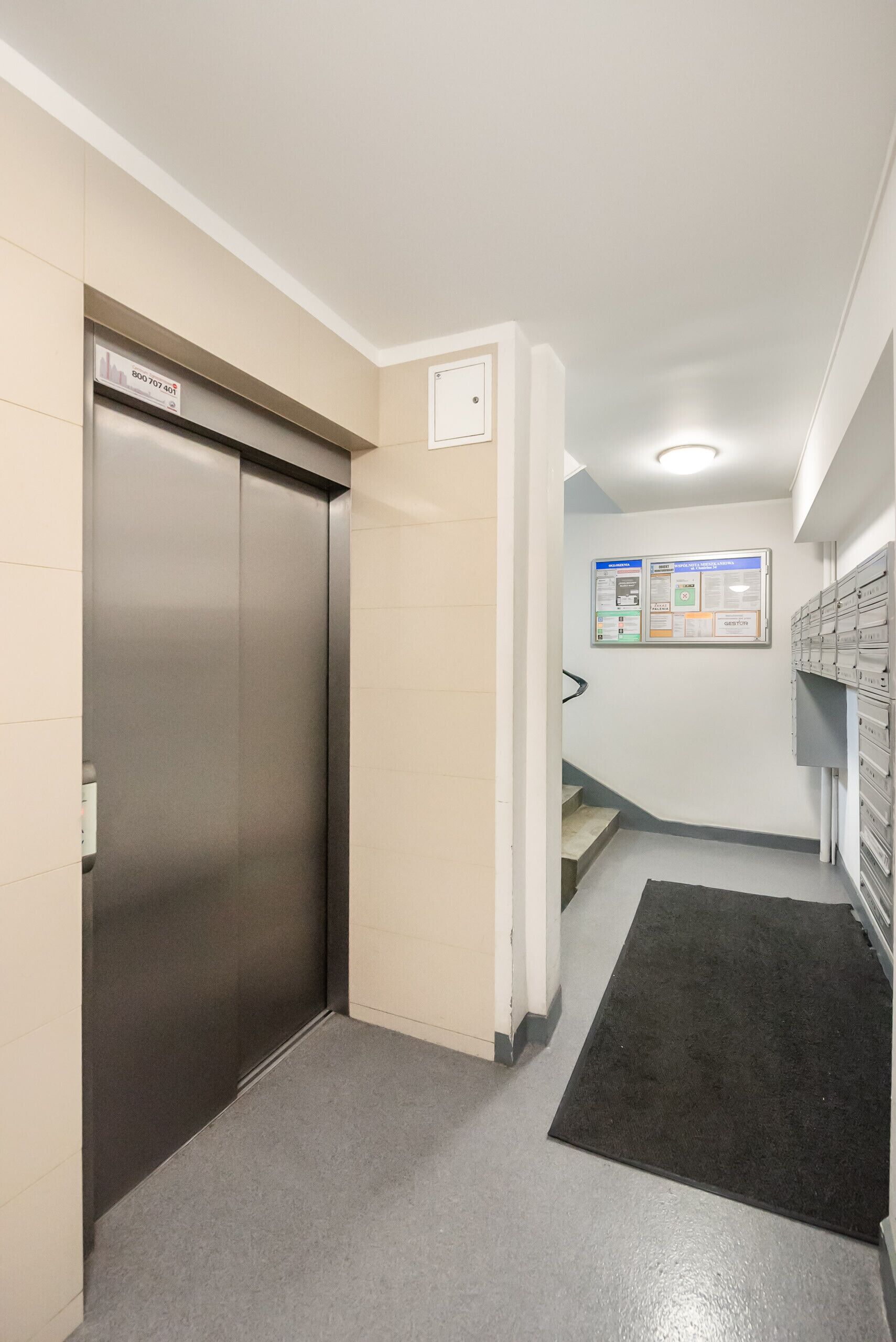 A hallway with a metallic elevator, a staircase, and a bulletin board with informational posters.