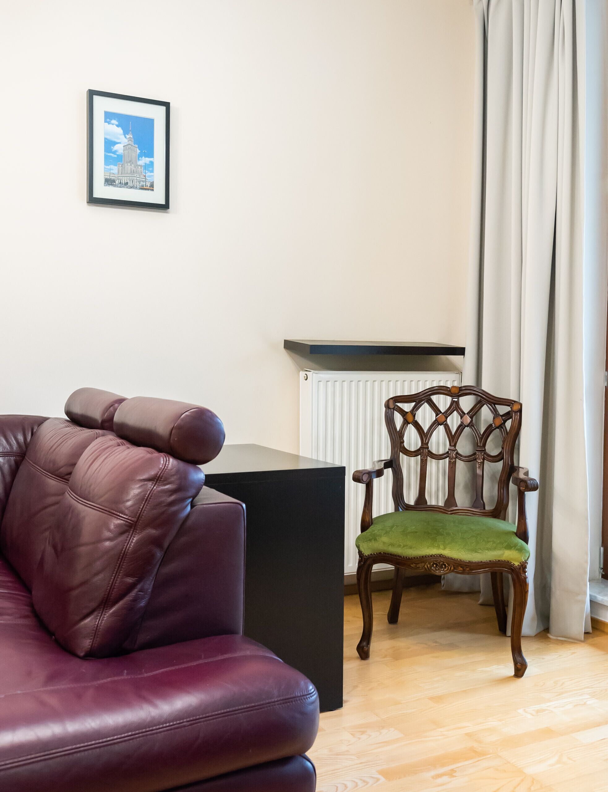 Un salon avec un canapé en cuir, une chaise en bois avec un coussin vert, une table d’appoint noire, un radiateur et un tableau encadré au mur.