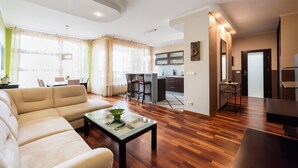 A modern living room with a sofa, coffee table, and dining area. There is a kitchen in the background with a bar and a TV.