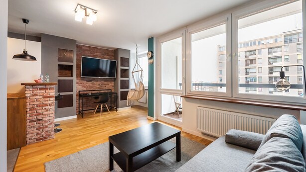 A modern living room with a TV, a brick accent wall, a hanging chair, and large windows. - Ratusz Arsenał Modern City Apartment (Warszawa)
