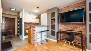A modern kitchen with a brick accent wall, a large TV, and a bar area with stools. - Ratusz Arsenał Modern City Apartment (Warszawa)