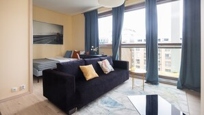 Een moderne woonkamer met een donkere bank, een bijzettafeltje en een bed met uitzicht op een stadslandschap.