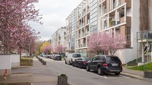 Een modern appartementencomplex met een rij geparkeerde auto's en kersenbloesems langs de straat.
