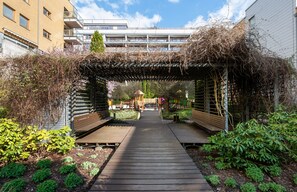 Een houten pad dat leidt naar een overdekt zitgedeelte met banken, omgeven door groen en een kleine tuin.