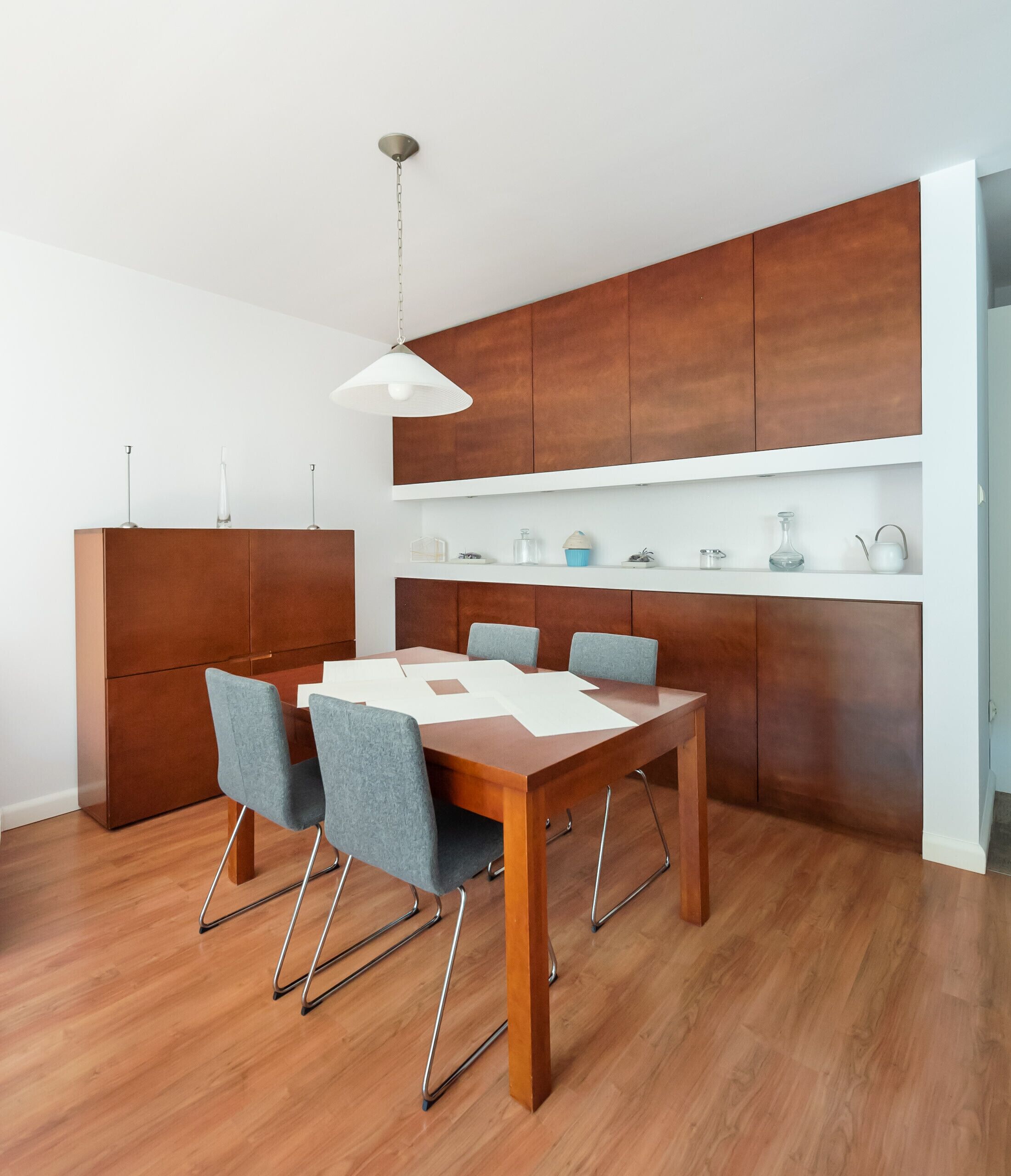 A modern dining area with a wooden table and chairs, a hanging light fixture, and built-in wooden cabinets above the table.