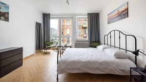 A bedroom with a bed, a dresser, a chair, and a view of buildings through large windows.