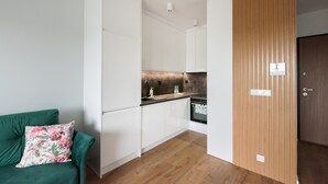 Une cuisine de taille réduite avec des meubles blancs, une porte en bois et un canapé vert agrémenté d’un coussin à motifs floraux.