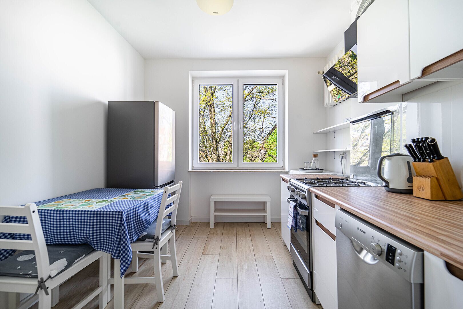 A compact kitchen with a dining table, a bench, a window with a view of trees, a stove, and a dishwasher.