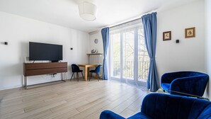 A modern living room with a flat-screen TV, wooden furniture, a dining area, and large windows with curtains.