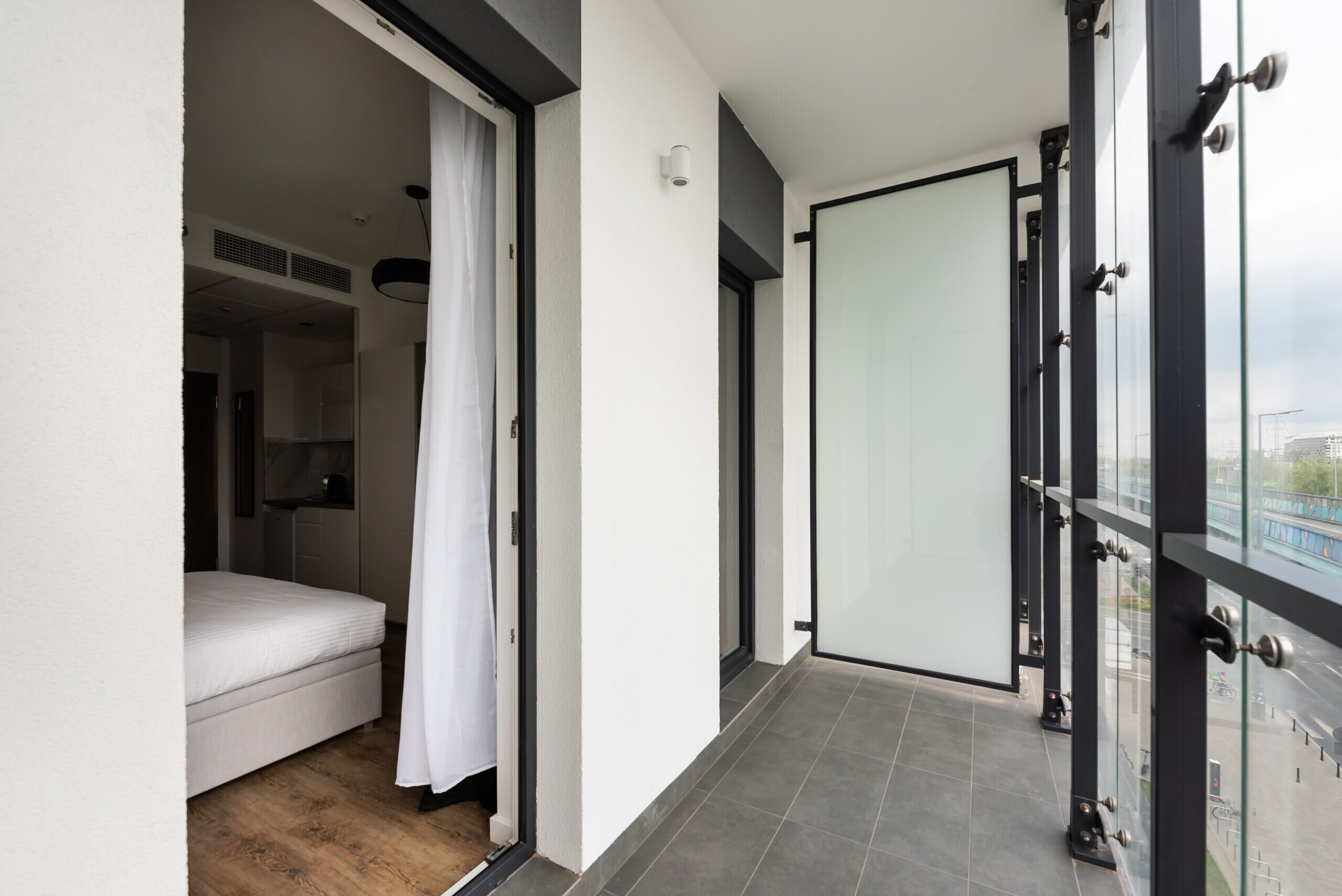 A modern hotel room with a sliding glass door leading to a balcony, a bed, and a white curtain.