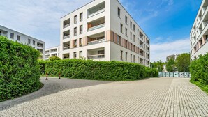 A modern multi-story building with balconies, a paved courtyard, and a hedge.