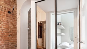 Una habitación con suelo de madera, pared de ladrillo y puerta corredera que da a un dormitorio con cama y mesita de noche.
