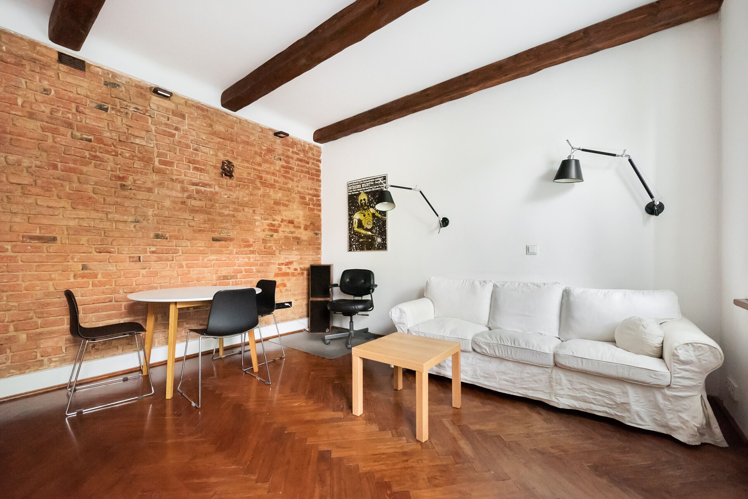 A living room with a white sofa, a wooden coffee table, and two black chairs. There is a brick wall and a white ceiling with exposed beams.