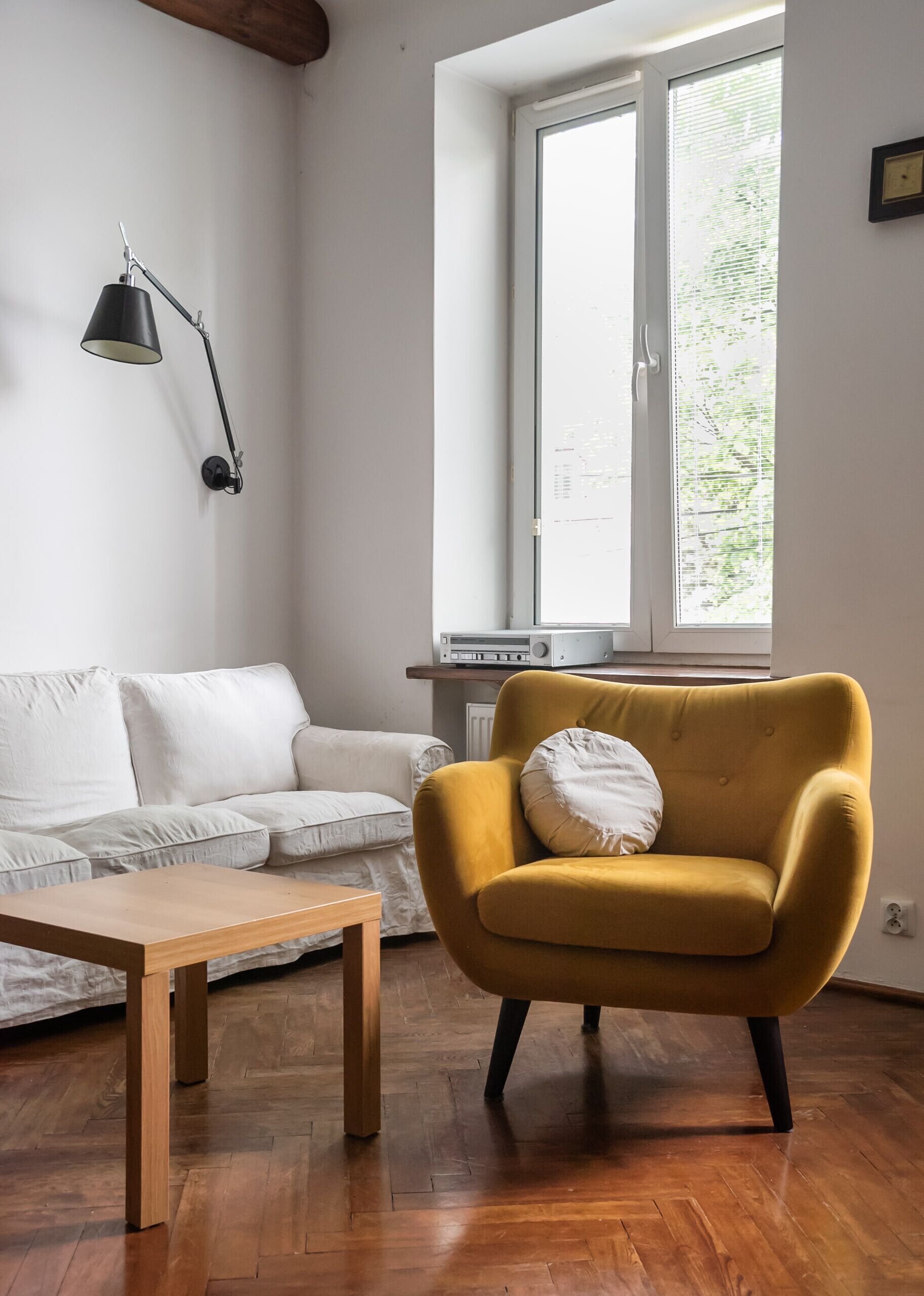 A living room with a white sofa, a mustard-colored armchair, a wooden coffee table, a black floor lamp, and a window with blinds.