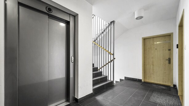 Un pasillo moderno con un ascensor metálico, una puerta de madera y una escalera con barandillas de metal.