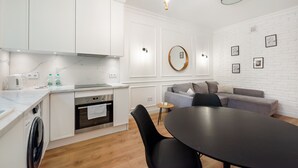 Une cuisine moderne avec des meubles blancs, un four encastré et un lave-linge. On y trouve également un coin repas avec une table et des chaises, ainsi qu’un canapé gris.
