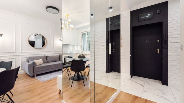 Un salon moderne avec un canapé gris, des chaises noires et une table à manger. Un grand miroir orne le mur, et une porte noire, dotée d’un trou de serrure, est également visible.