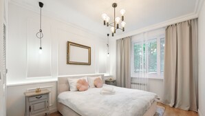 Une chambre à coucher avec un grand lit, des tables de chevet, un lustre, un tableau encadré et une fenêtre avec des rideaux.