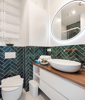 A modern bathroom with a white toilet, a wooden vanity, a large round mirror, and a herringbone-patterned backsplash. - Wola Kasprzaka Luxury Apartment (Warszawa)