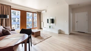A modern living room with a wooden floor, a dining table, a sofa, a TV mounted on the wall, and large windows with curtains. - Magazynowa Classy Apartment (Warszawa)