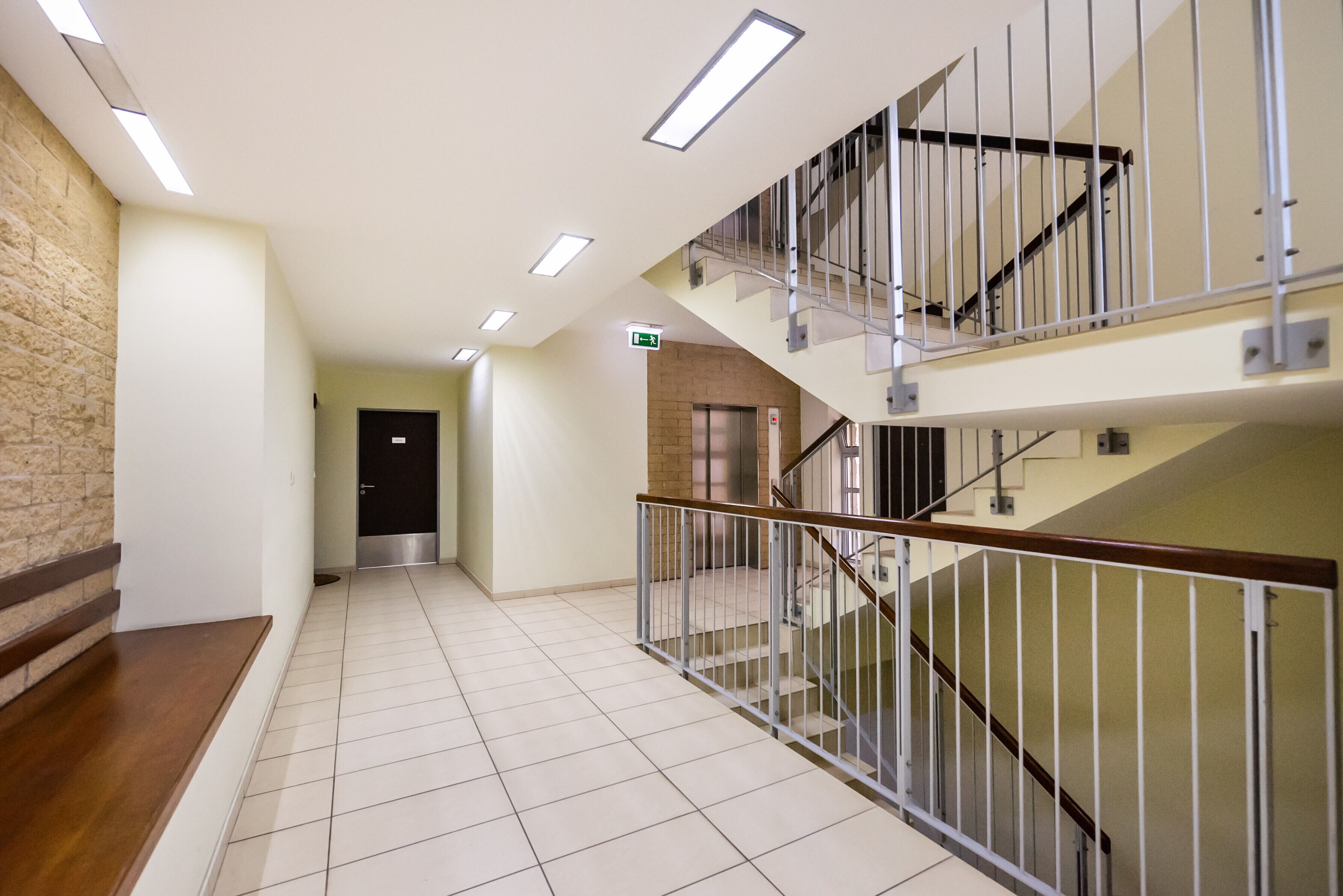 Un pasillo de hotel con suelo de baldosas, un banco de madera, una escalera con barandillas de metal y un ascensor.