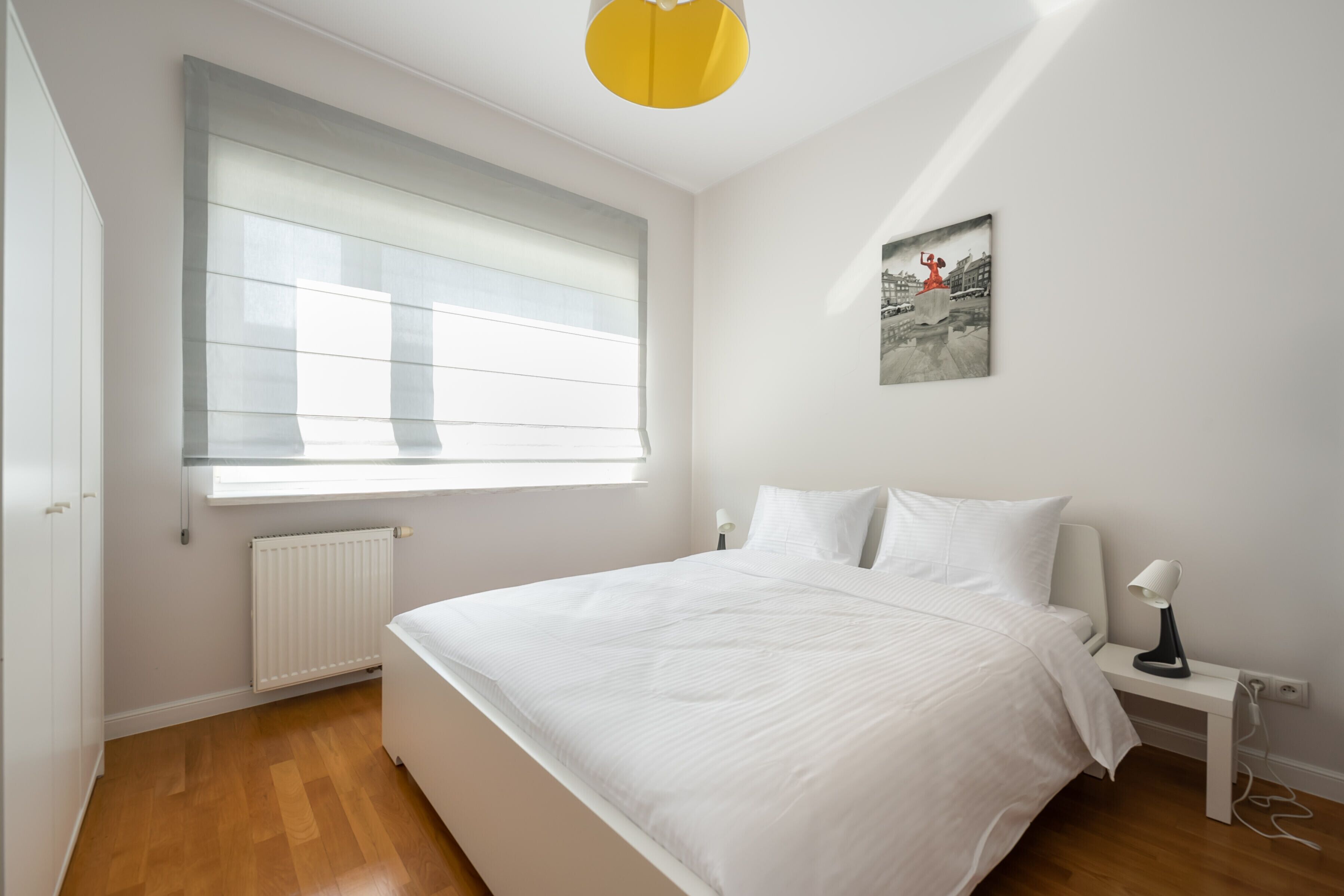 A bedroom with a large bed, a bedside table with a lamp, a window with blinds, a radiator, and a wall-mounted picture.