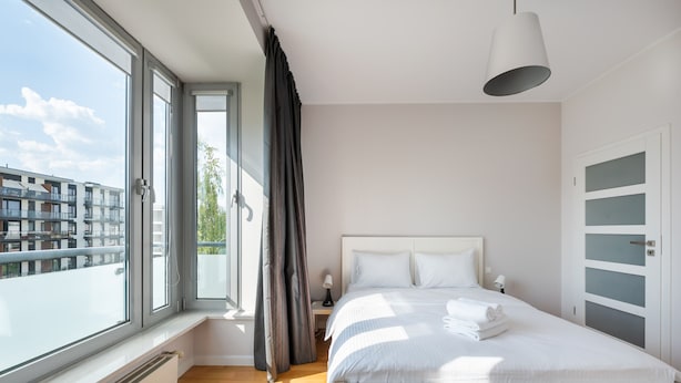 A modern bedroom with a large window, a bed with white linens, a bedside table with a lamp, and a view of buildings outside. - Mokotów Serenity Premium Apartment (Warszawa)