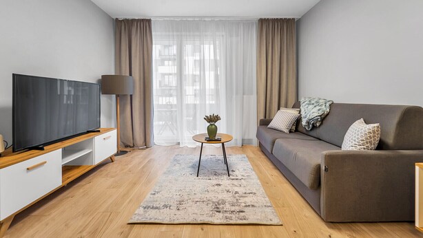 Ein modernes Wohnzimmer mit einem Flachbildfernseher auf einem Holzboden, einem grauen Sofa mit gemusterten Kissen, einem runden Couchtisch mit einer Vase und einem großen Fenster mit durchsichtigen Vorhängen.