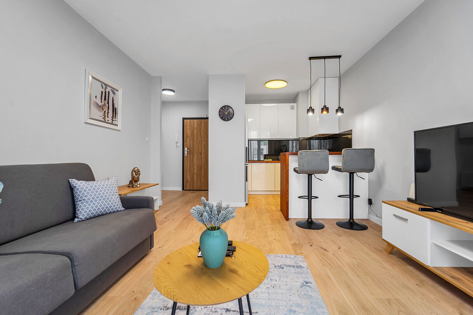 A modern living room with a grey sofa, a coffee table, and a TV. There is a kitchen in the background with white cabinets and a wooden countertop.