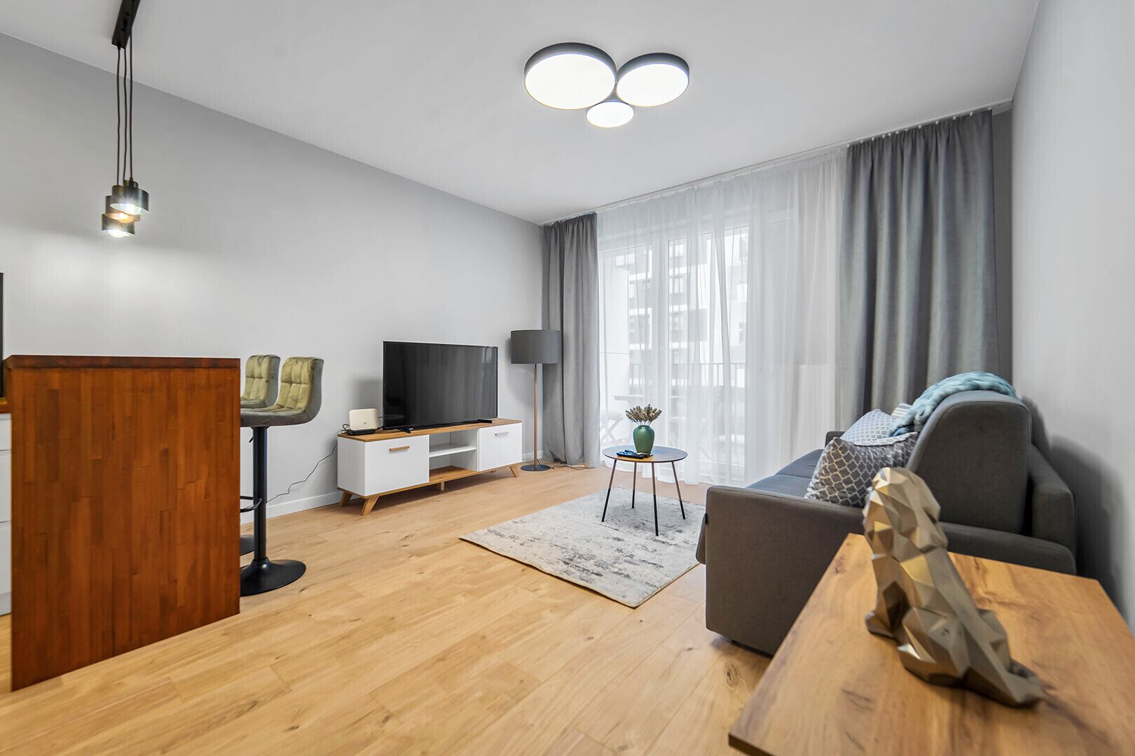 Una sala de estar moderna con piso de madera, un televisor de pantalla plana sobre un mueble blanco, un sofá gris, una mesa de centro de madera y una lámpara de pie negra.